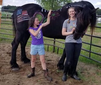 Two girls painting an American flag and designs onto the side of a tall black horse