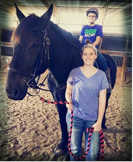 A smiling young woman holding a red and black lead rope beside a large dark horse, with a young rider in a helmet on its back